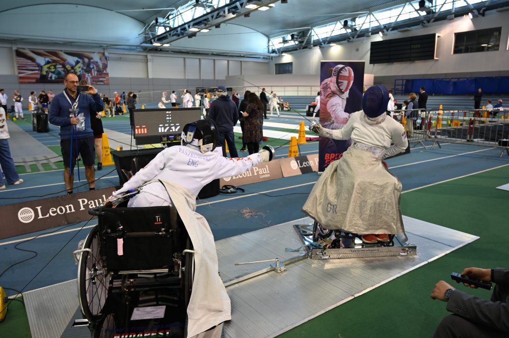 two wheelchair users fencing at a competition strapped into a shiny new fencing frame