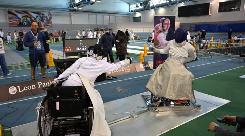 two wheelchair users fencing at a competition strapped into a shiny new fencing frame