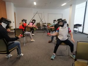 People sitting in chairs fencing against each other using plastic equipment. 