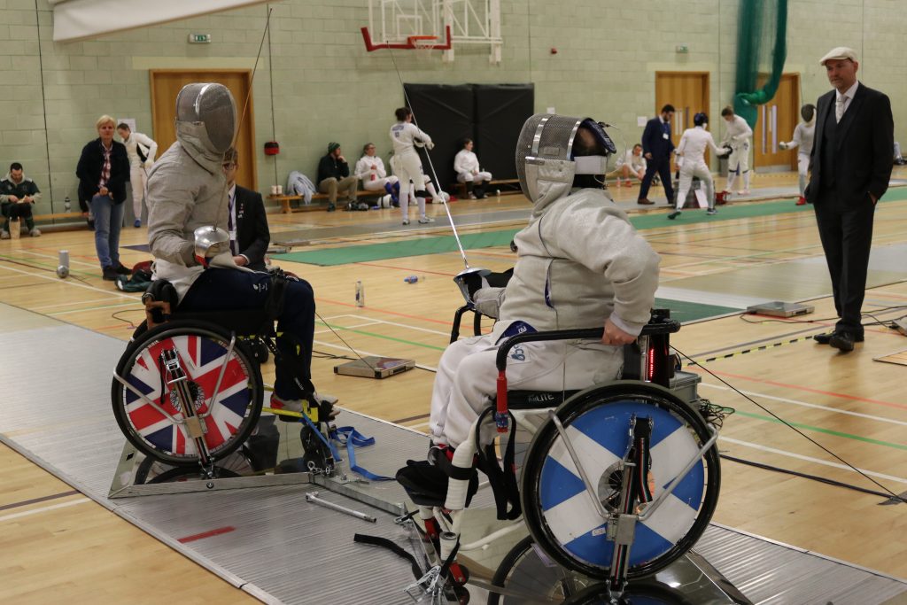 Two wheelchair fencers facing off against each other, one has the union flag as their wheel cover and the other has the Saltire flag of Scotland