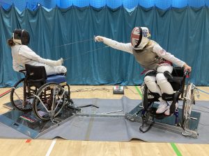 Two wheelchair using fencers face off one is clearly on the attack sword thrusting foward while the other leans back to dodge and raises their weapon in defence. 