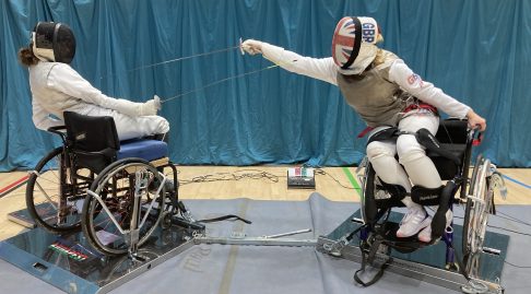 Two wheelchair using fencers face off one is clearly on the attack sword thrusting foward while the other leans back to dodge and raises their weapon in defence.