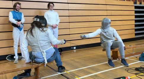 Two fencers facing off sitting on benches with tow other refereeing the bout.
