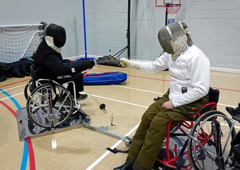 Two wheelchair fencers facing off in a Sports Center club environment, their chairs locked into a wheelchair fencing frame. 