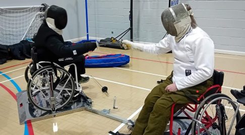 Two wheelchair fencers facing off in a Sports Center club environment, their chairs locked into a wheelchair fencing frame.