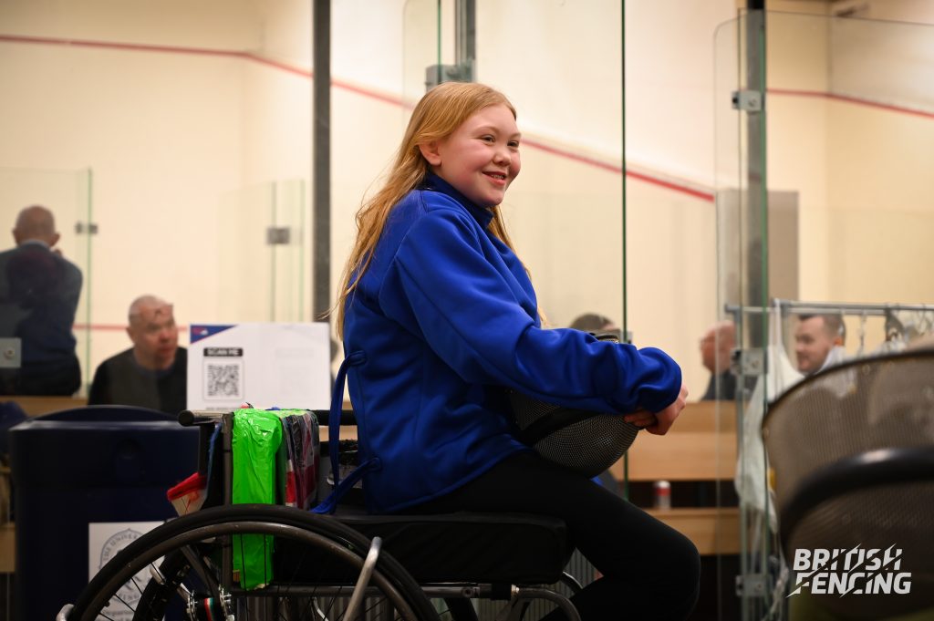 A young girl sitting on a wheelchair smiling, she is in a fencing jacket and holding a fencing mask in her hands. 