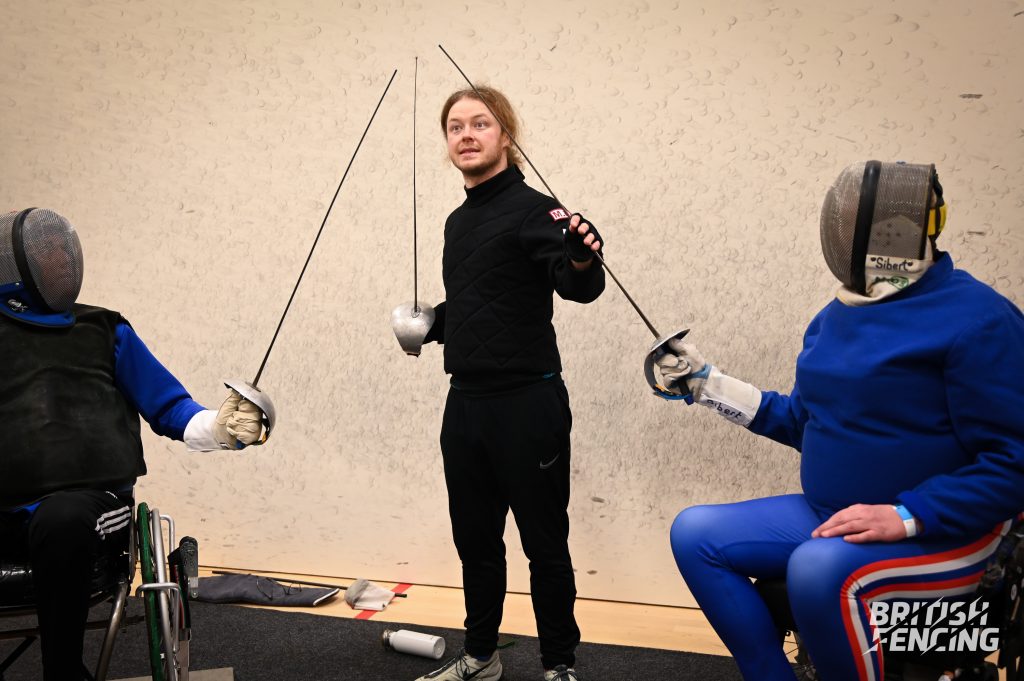 A male fencing coach dressed all in black holds a sword standing between two seated fencers also holding swords (making a triangular shape) 