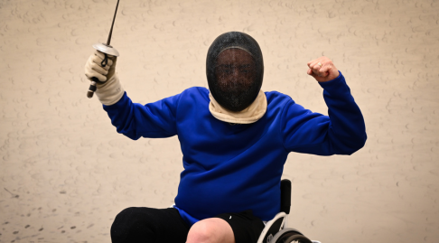 A participant in a fencing jacket and mask, sitting with sword in hand holding a triumphant pose looks excited to be participating.