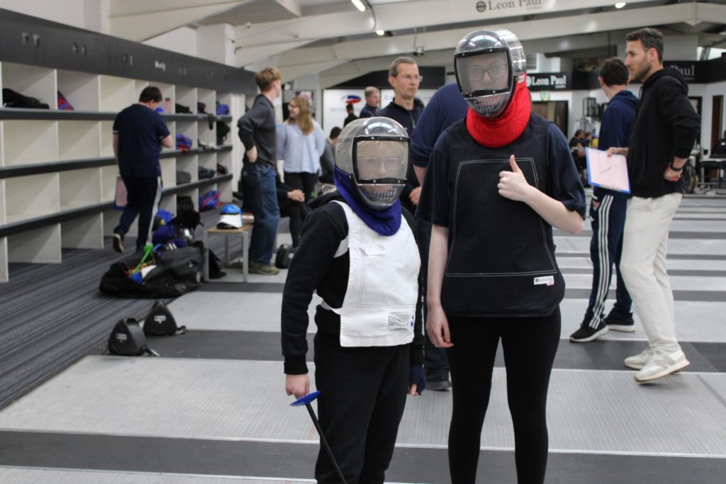 two young people in plastic fencing masks and bibs, facing the camera, one has their thumb up. 