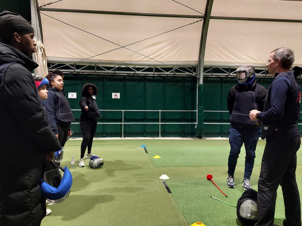 Several people are taking part in a fencing training activity with plastic swords and masks. A coach is talking to them.