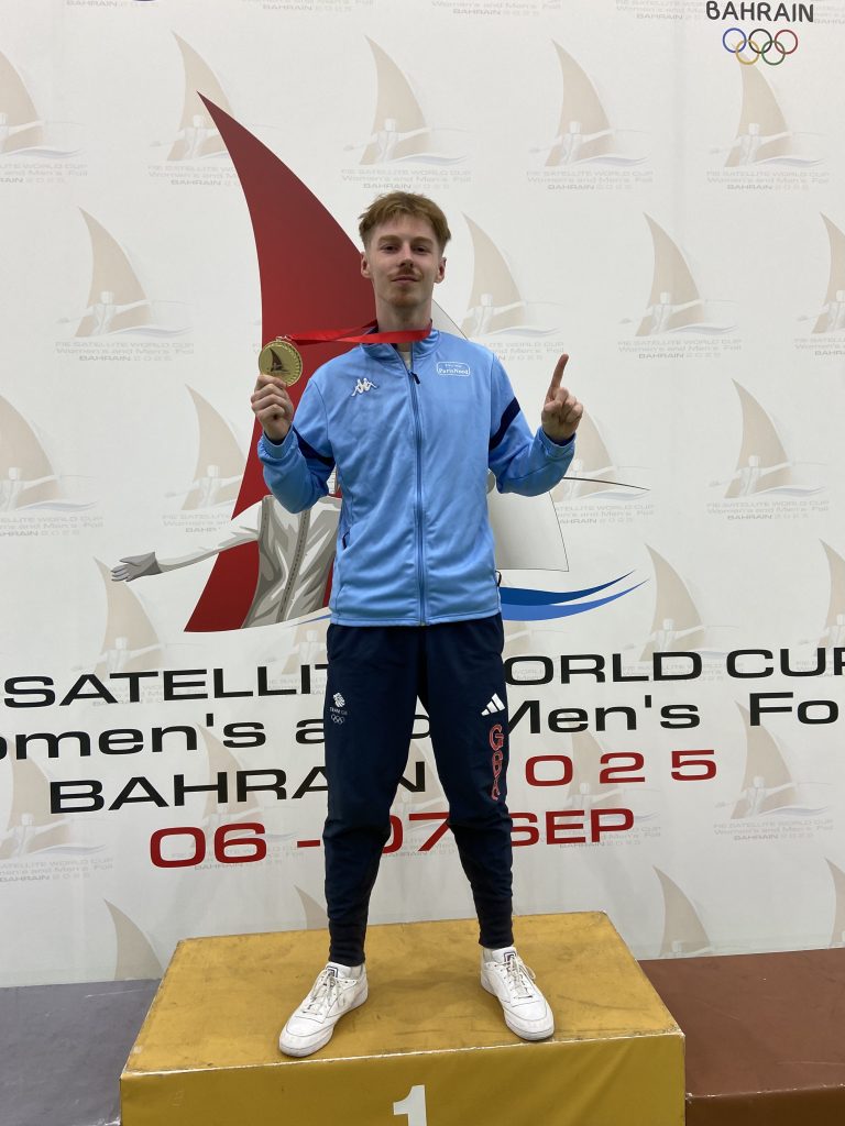 A fencer stands on a podium holding up a gold medal.