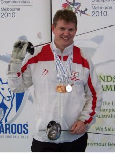 Paul Abrahams on a podium with a medal around his neck, smiling to camera, holding fencing swords in his hands
