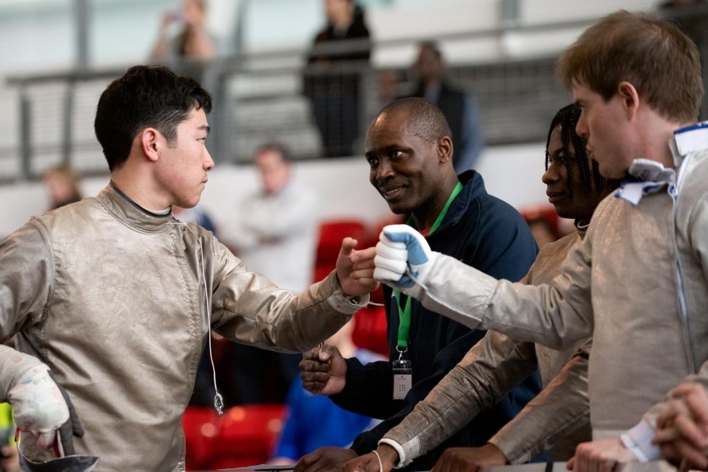 Two sabre fencers fist pump next to a coach who is smiling at them, as well as another fencer.
