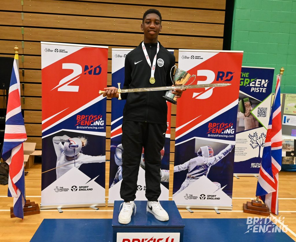 A fencer stands on a podium smiling, while holding a trophy and ceremonial sword.