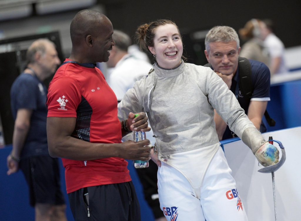 A coach and sabre fencer are talking and smiling.