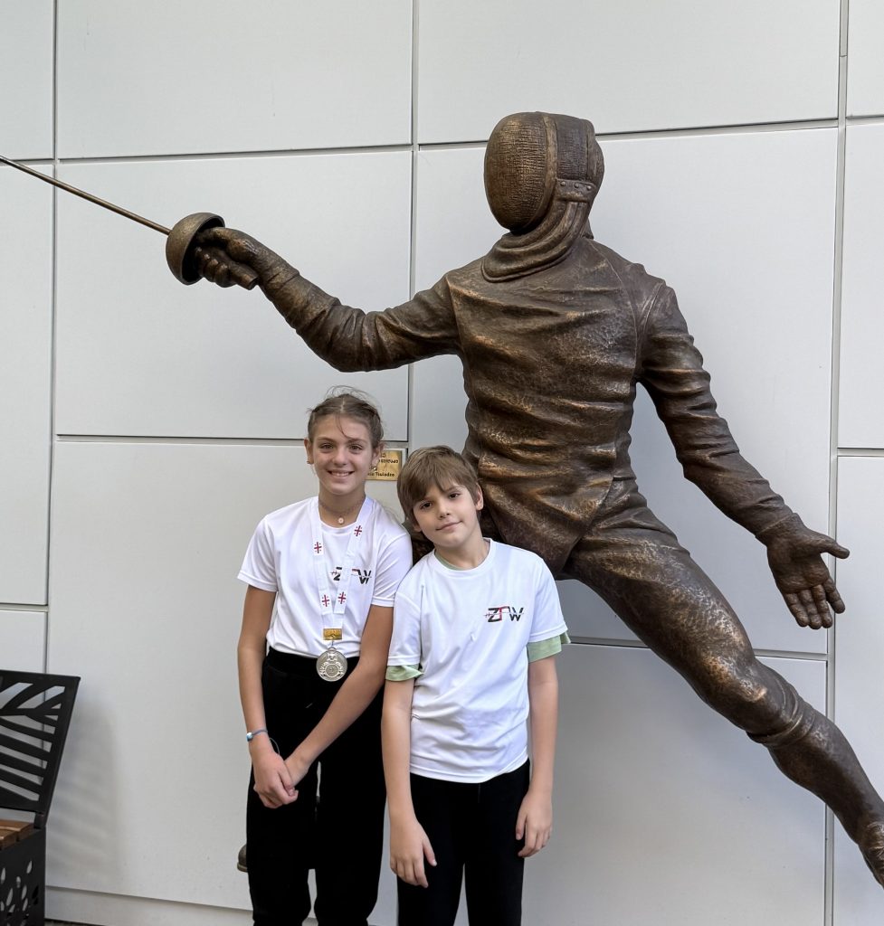 Two young fencers standing in front of a fencing statue smile to the camera.