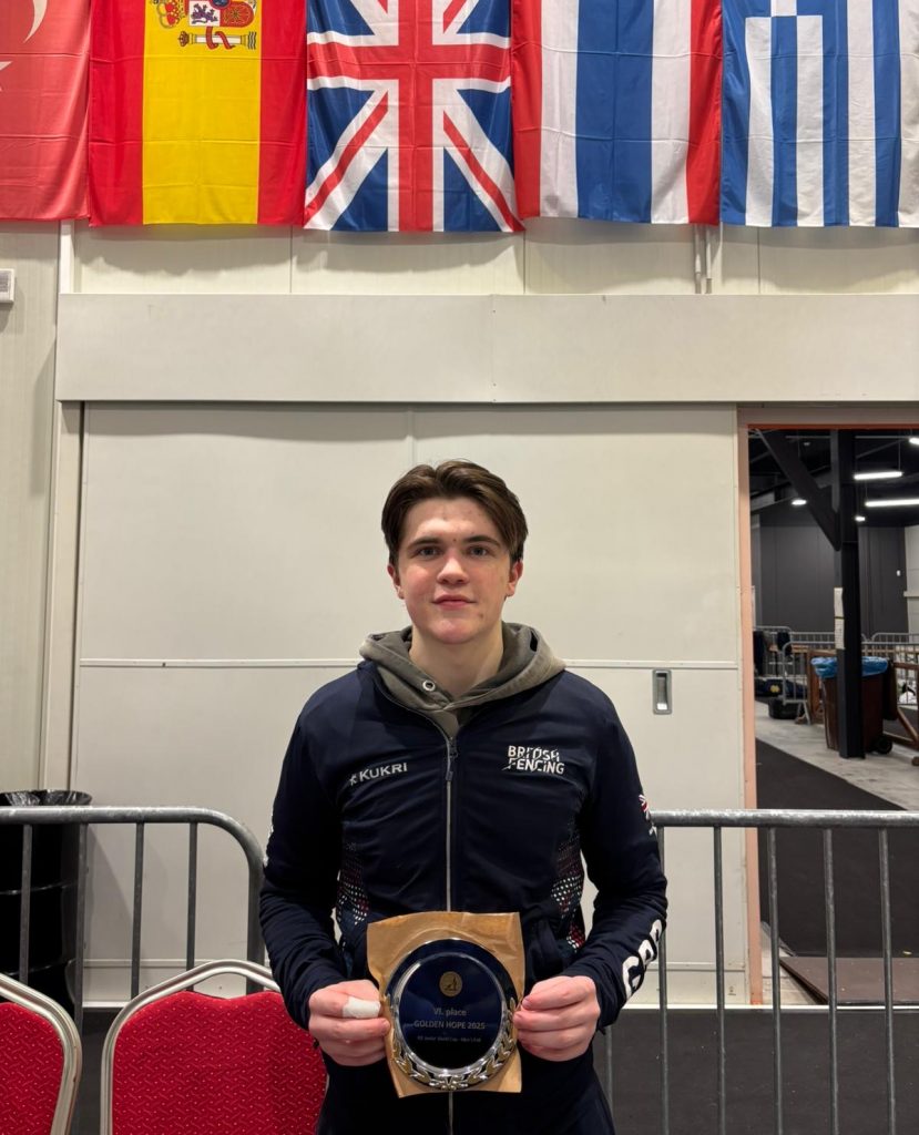 A fencer smiles to the camera holding a trophy plate.
