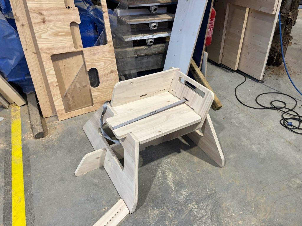 A wooden SwordSeat with a grey strap on a concrete floor of some sort of cutting facroty, based on the equipment seen in the background. 