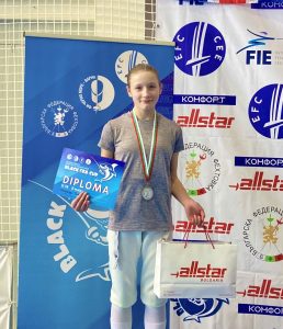 A fencer on a podium holding a certificate and trophy smiles to the camera.