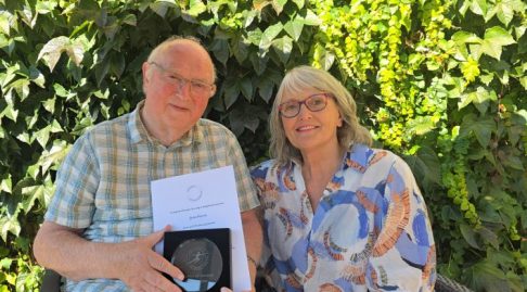 John Mason smiling with Gillian Aghajan, who has presented him with a certificate and award he is holding showing him as Member of Honour of EVF. This was on 25th August.