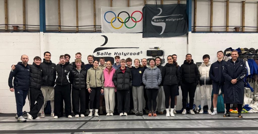 A large group of people in a fencing salle smile to the camera.