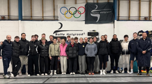 A large group of people in a fencing salle smile to the camera.