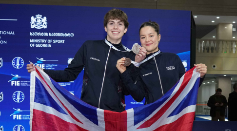 Two GBR fencers smile to the camera, holding up bronze medals and the GBR flag.