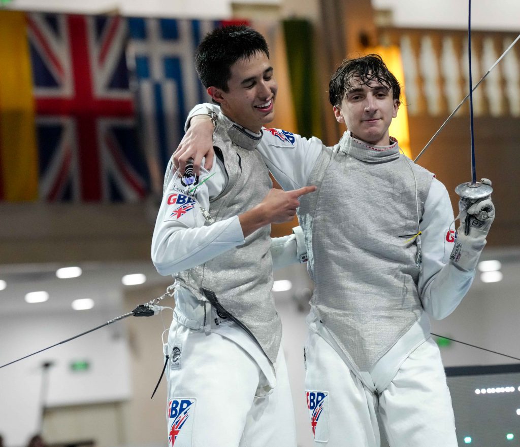 Two GBR foil fencers smile to the camera, one pointing at the other