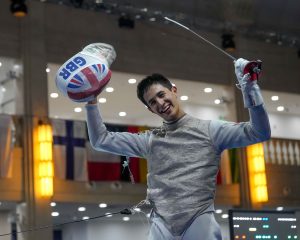 A GBR foil fencer celebrates his victory, holding up his mask and sword.