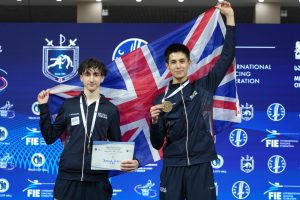 Two GBR fencers holding up medals and certificates smile to the camera, holding up the GBR flag.