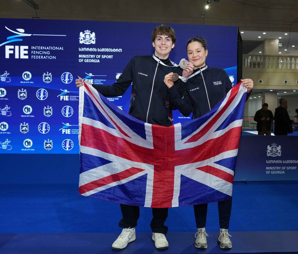 Two GBR fencers smile to the camera, holding up bronze medals and the GBR flag.