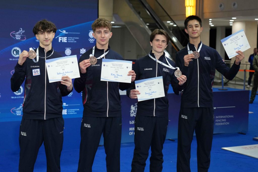 Four GBR fencers smile to the camera, holding up bronze medals and certificates