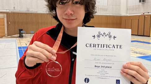 A young fencer smiles to the camera, holding up a medal and certificate.