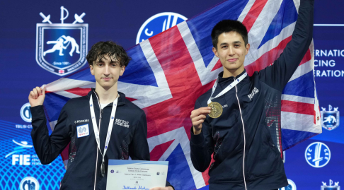 Two GBR fencers holding up medals and certificates smile to the camera, holding up the GBR flag.