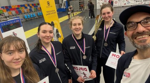 A selfie of four GBR foil fencers smiling while holding up certificates and medals, alongside their coach. One is holding a trophy.