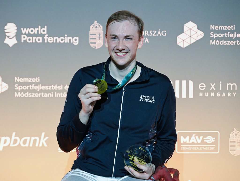 A GBR para fencer smiles to the camera, holding up a gold medal and trophy.
