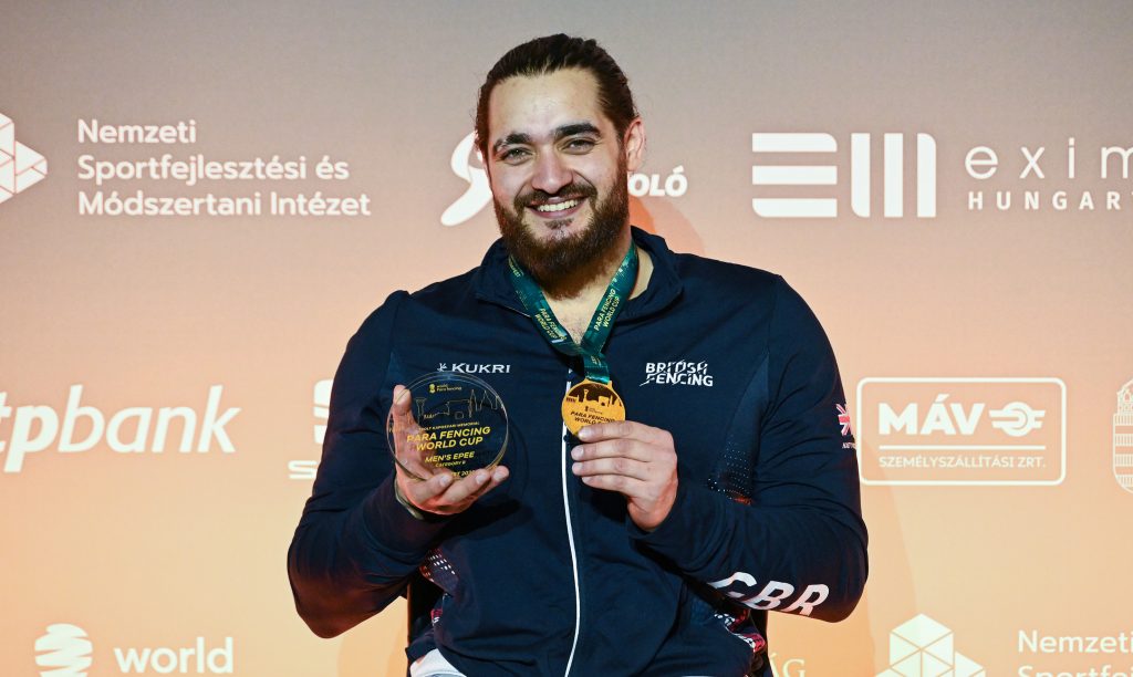 A GBR para fencer smiles to the camera, holding up a gold medal and trophy.