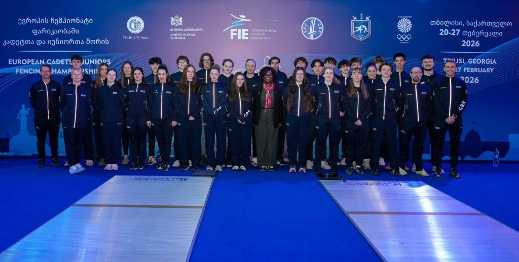 A large group of Junior fencers, coaches and support team smile to the camera.