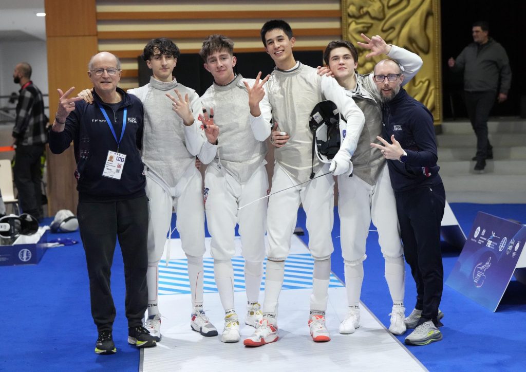 Four GBR foil fencers and two coaches smile to the camera, holding up three fingers each