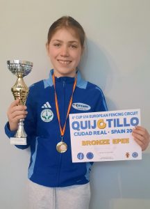 A young fencer holding up a certificate, trophy and wearing a medal smiles to the camera