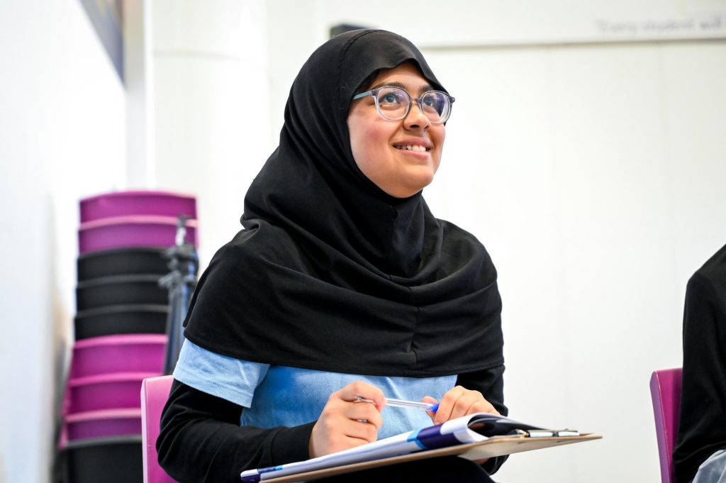 A young person in a hijab looks up and smiles while writing in a workbook.