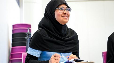 A young person in a hijab looks up and smiles while writing in a workbook.