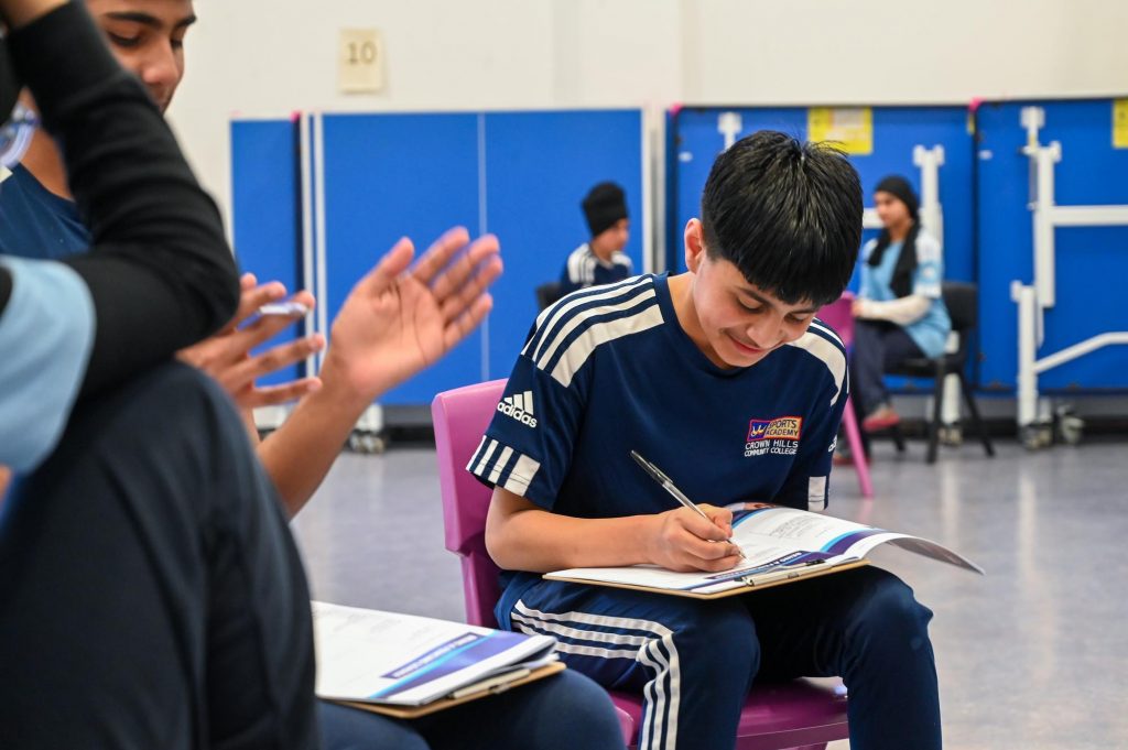 A Crown Hills student writes in their workbook. Several other students are in the background.