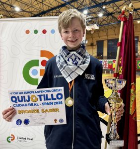 A young fencer holding up a certificate, trophy and wearing a medal smiles to the camera