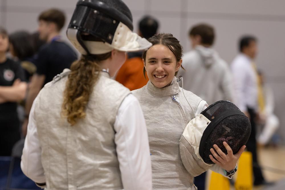 Two young foil fencers are facing each other, and one is smiling to the other.