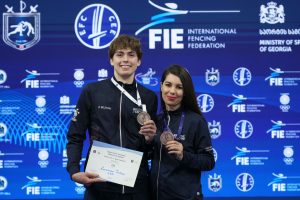A fencer and their coach, both wearing bronze medals, smile to the camera