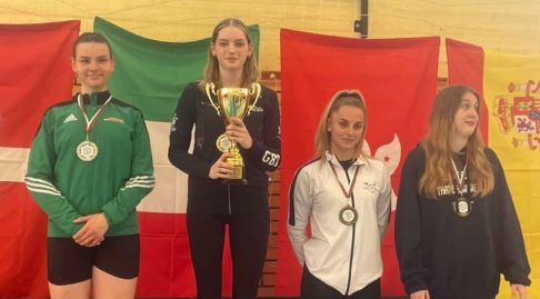 Four fencers stand on a podium smiling to the camera. One is holding a trophy