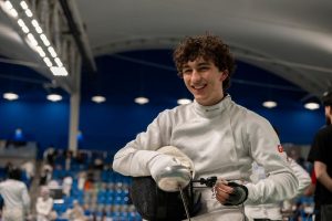 An epee para fencer at a competition holds his mask and sword while smiling