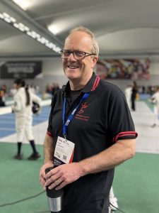 A fencing coach smiles to the camera. He is at the BYCs in Sheffield, with fencers competing in the background and other people watching.