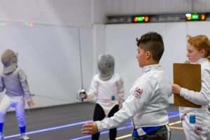 Two young sabre fencers are training at a club. Another is refereeing and another is holding a clipboard.